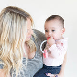 A woman with long blonde hair holds a baby in a pink shirt chewing on a Bumkins Silicone Teething Charms: Pink.