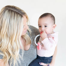 Load image into Gallery viewer, A woman with long blonde hair holds a baby in a pink shirt chewing on a Bumkins Silicone Teething Charms: Pink.