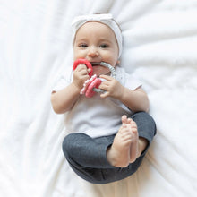 Load image into Gallery viewer, A baby wearing a bow headband on a white blanket, smiling softly, chews a Bumkins Silicone Teething Charms: Pink.