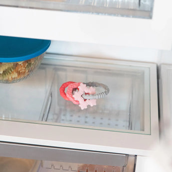 Close-up of a glass refrigerator shelf with a Bumkins Silicone Teething Charms: Pink beside a covered bowl of food.