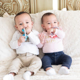 Two babies on a beige blanket chew teething toys, including a Bumkins Silicone Teething Charms: Pink.