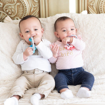 Two babies on a beige blanket chew teething toys, including a Bumkins Silicone Teething Charms: Pink.