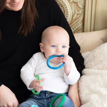 Load image into Gallery viewer, A baby in a white shirt holds and chews Bumkins Silicone Teething Rings 4 Pack: Summer, sitting on an adult's lap in a cozy room.