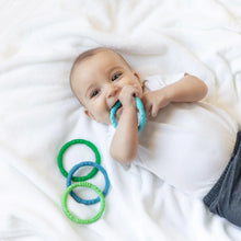 Load image into Gallery viewer, A baby on a white blanket chews a Bumkins Silicone Teething Rings 4 Pack: Summer, while the others sit nearby.