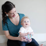 A woman smiles at a blue-eyed baby holding Bumkins Silicone Teething Rings 4 Pack: Spring, both on the floor against a light wall.