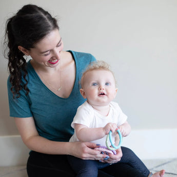 A woman smiles at a blue-eyed baby holding Bumkins Silicone Teething Rings 4 Pack: Spring, both on the floor against a light wall.