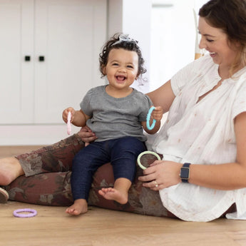 A child sits on a woman's lap on a wood floor, smiling while holding Bumkins Silicone Teething Rings 4 Pack: Spring.