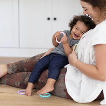 Load image into Gallery viewer, A woman sits with a laughing toddler holding Bumkins Silicone Teething Rings 4 Pack: Spring on her lap on a wood floor in a cozy home.
