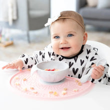 Load image into Gallery viewer, A baby in a bow sits at a white table with a gray bowl and spoon on a pink placemat, wearing a Bumkins Sleeved Bib: Hearts.