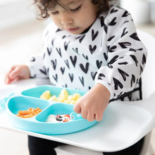 Load image into Gallery viewer, A toddler in a Bumkins Sleeved Bib: Hearts sits in a high chair, eating yogurt, fruit, & eggs from a blue divided plate.