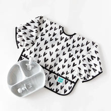 Load image into Gallery viewer, Bumkins Sleeved Bib: Hearts lays next to a gray divided plate and toddler utensils on a white background.