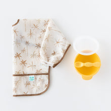 Load image into Gallery viewer, Bumkins Sleeved Bib: Palm Daze sits folded beside a yellow bowl and spoon on a white backdrop.