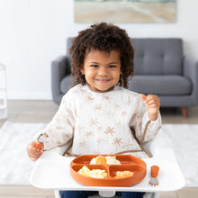Load image into Gallery viewer, In a high chair, a curly-haired child wearing a Bumkins Sleeved Bib: Palm Daze smiles while holding a spoon next to a divided plate of food.