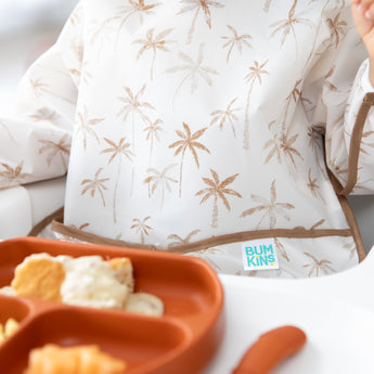 Close-up of a child in a Bumkins Sleeved Bib: Palm Daze enjoying baby-led weaning with an orange plate of crackers and dip in the foreground.