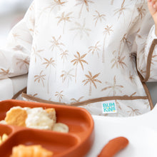 Load image into Gallery viewer, Close-up of a child in a Bumkins Sleeved Bib: Palm Daze enjoying baby-led weaning with an orange plate of crackers and dip in the foreground.