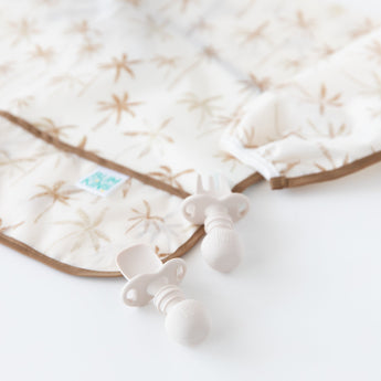 Close-up of Bumkins Sleeved Bib: Palm Daze next to two beige silicone utensils on a white background.