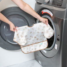 Load image into Gallery viewer, A person holds Bumkins Sleeved Bib: Palm Daze in front of an open front-loading washing machine.