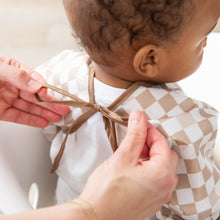 Load image into Gallery viewer, An adult ties the brown ribbon closure on a Bumkins Sleeved Bib: Sand Check as a baby in a white shirt sits ready for baby-led weaning.