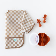Load image into Gallery viewer, Bumkins Sleeved Bib: Sand Check lays folded next to an orange bowl & spoon set and orange toddler utensils on a white background.