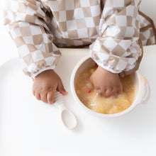 Load image into Gallery viewer, A child in a Bumkins Sleeved Bib: Sand Check joyfully explores mushy food on a high chair tray, with a spoon nearby, embracing baby-led weaning.