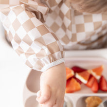 Load image into Gallery viewer, Close-up of a child in a Bumkins Sleeved Bib: Sand Check reaching for strawberries and oranges, embracing baby-led weaning.