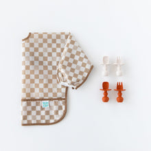 Load image into Gallery viewer, Bumkins Sleeved Bib: Sand Check lays folded next to two sets of silicone utensils on a white background.