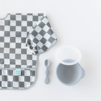 Bumkins Sleeved Bib: Charcoal Check lays next to a gray bowl and spoon on a white background.