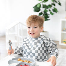 Load image into Gallery viewer, A child in a Bumkins Sleeved Bib: Charcoal Check sits in a high chair, smiling with a spoon, ready for baby-led weaning fun.