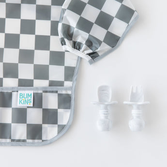 Close-up of Bumkins Sleeved Bib: Charcoal Check next to baby utensils on a white background, ideal for baby-led weaning.