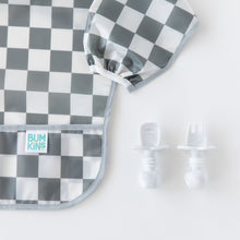 Load image into Gallery viewer, Close-up of Bumkins Sleeved Bib: Charcoal Check next to baby utensils on a white background, ideal for baby-led weaning.