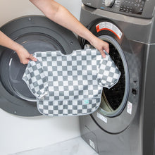 Load image into Gallery viewer, A person holds Bumkins Sleeved Bib: Charcoal Check in front of an open front-loading washing machine.