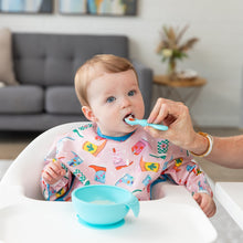 Load image into Gallery viewer, A baby in a high chair wears a Bumkins Sleeved Bib: Boots, being spoon-fed. A blue bowl sits on the tray with a sofa and plant behind.