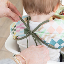 Load image into Gallery viewer, Adult hands tie the ribbon closure of a Bumkins Sleeved Bib: Motley Check around a child's neck.