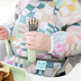 Close-up of a child in a colorful checkered sleeved bib holding Bumkins Spoon + Fork: Sage.