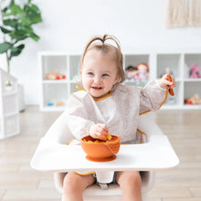 Load image into Gallery viewer, A toddler wearing a Bumkins Sleeved Bib: Wander smiles while eating from an orange bowl at a high chair; toys fill shelves behind.