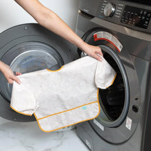 Load image into Gallery viewer, A person holding a Bumkins Sleeved Bib: Wander in front of an open front-loading washing machine.