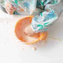 Load image into Gallery viewer, Close-up of a child's hand playing with yogurt in an orange bowl, wearing a Bumkins Sleeved Bib: Dinosaurs.