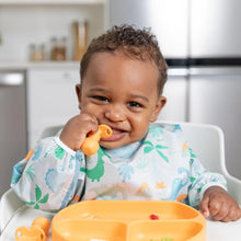Load image into Gallery viewer, A baby in a Bumkins Sleeved Bib: Dinosaurs sits happily in a high chair with a spoon and orange plate.