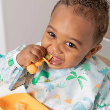 Load image into Gallery viewer, A smiling baby joyfully eats with an orange spoon and plate, wearing a Bumkins Sleeved Bib: Dinosaurs.