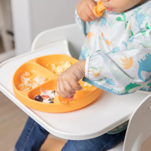 Load image into Gallery viewer, Close-up of a toddler in a high chair wearing a Bumkins Sleeved Bib: Dinosaurs, eating from an orange divided plate with a spoon.
