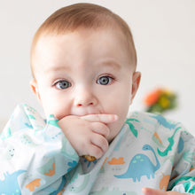 Load image into Gallery viewer, A baby with big eyes wears a Bumkins Sleeved Bib: Dinosaurs while sucking their thumb.