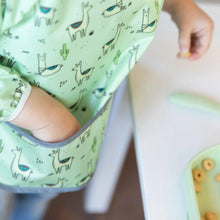 Load image into Gallery viewer, Close-up of a child reaching into the pocket of a Bumkins Sleeved Bib: Llamas near a white table with cereal pieces on a green plate.