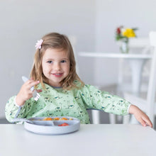 Load image into Gallery viewer, A girl in a Bumkins Sleeved Bib: Llamas and pink flower clip smiles while holding a fork over a gray divided plate on a white table.