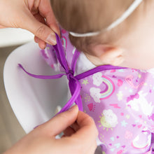 Load image into Gallery viewer, An adult ties the ribbon closure of a Bumkins Sleeved Bib: Unicorns around a child's neck.