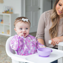 Load image into Gallery viewer, A baby in a high chair wears a Bumkins Sleeved Bib: Unicorns, enjoying self-feeding from a purple bowl while an adult nearby smiles warmly.