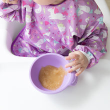 Load image into Gallery viewer, A baby in a high chair wearing a Bumkins Sleeved Bib: Unicorns eagerly holds a lavender bowl of applesauce.