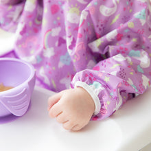 Load image into Gallery viewer, A child wears a Bumkins Sleeved Bib: Unicorns while sitting at a table, holding a lavender bowl filled with food.