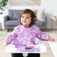 Load image into Gallery viewer, A child in a Bumkins Sleeved Bib: Unicorns smiles in a high chair, enjoying food from a purple divided plate with matching utensils.