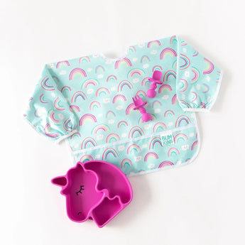 Bumkins Sleeved Bib: Rainbows lays on a white background with a dark pink unicorn-shaped plate and matching utensils.