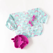 Load image into Gallery viewer, Bumkins Sleeved Bib: Rainbows lays on a white background with a dark pink unicorn-shaped plate and matching utensils.
